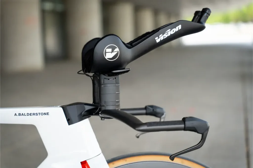 Primer plano de la parte delantera de una bicicleta de contrarreloj blanca, con manillar Vision aerodinámico y extensiones, con el freno delantero expuesto -igual que los utilizados por Las cadenas KMC impulsan al Caja Rural-Seguros RGA- sobre un fondo urbano difuminado.