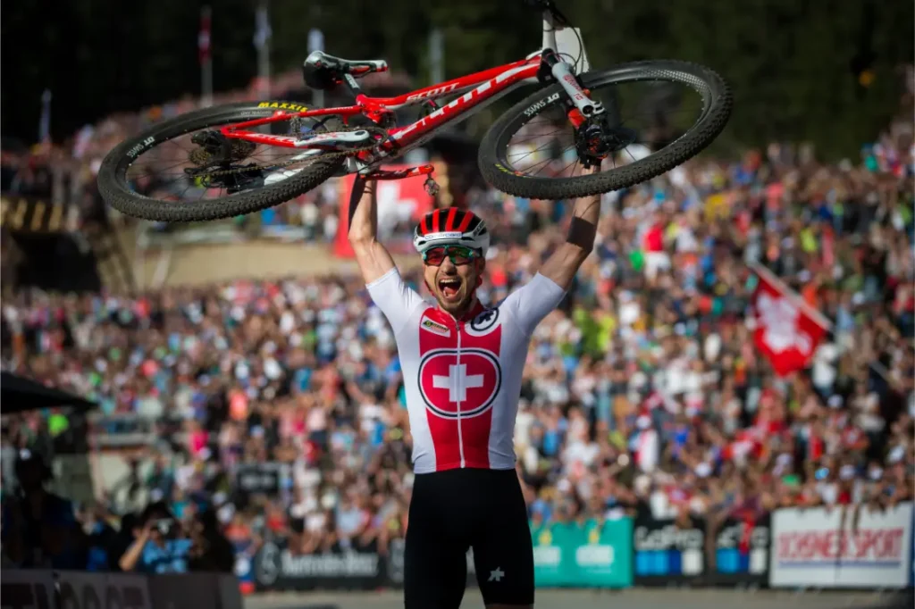 Un ciclista con maillot suizo levanta una bicicleta de montaña en alto, celebrando la retirada de Nino Schurter ante una gran multitud en un acto al aire libre.