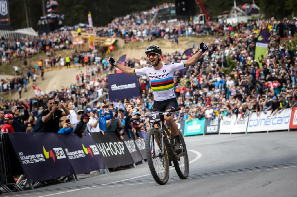 Un ciclista cruza la línea de meta con los brazos en alto en señal de victoria mientras una gran multitud le aclama en una carrera profesional de bicicleta de montaña, en honor a la retirada del legendario Nino Schurter.