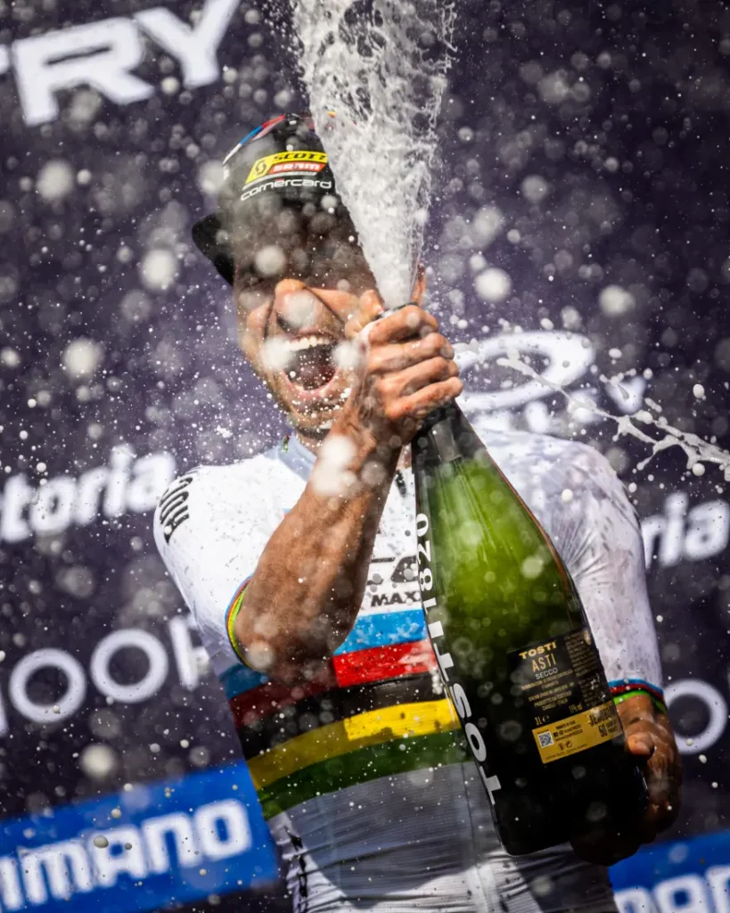 Un ciclista con un maillot de rayas arco iris rocía champán con una amplia sonrisa en la etapa, celebrando la retirada de Nino Schurter mientras las gotas y la espuma llenan el aire.
