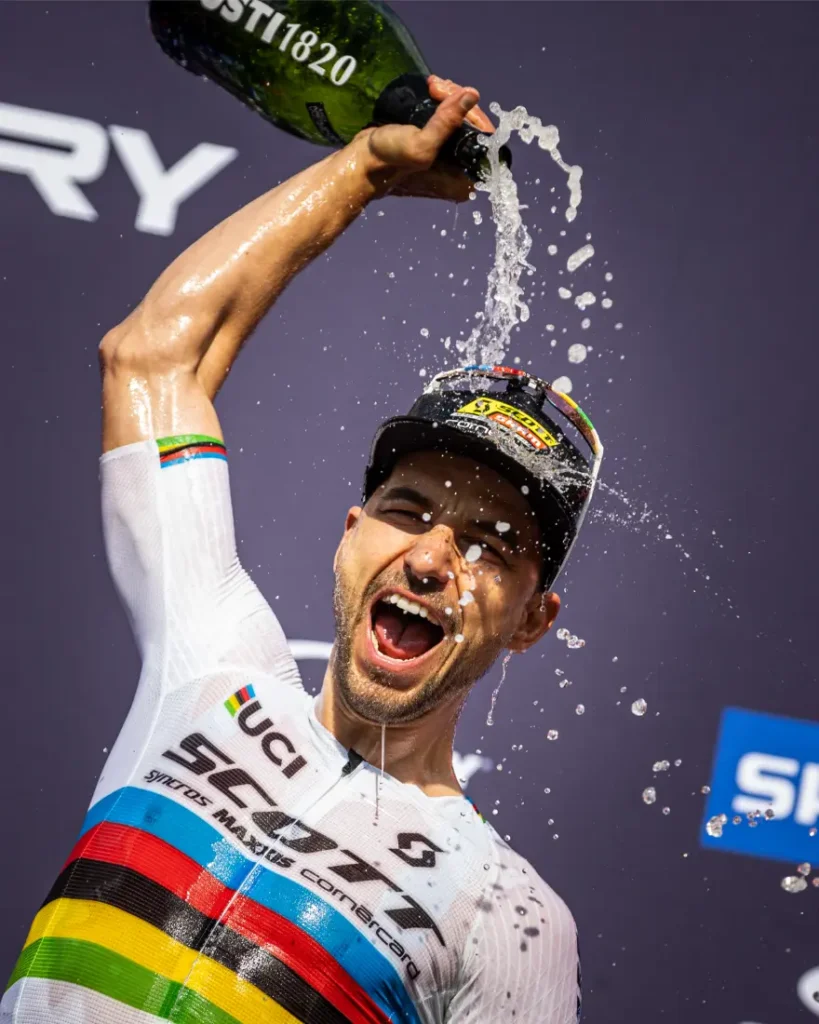 Un ciclista con maillot arco iris, celebrando la retirada como Nino Schurter, se echa champán por la cabeza, con la boca abierta, sobre un fondo oscuro de acontecimientos.