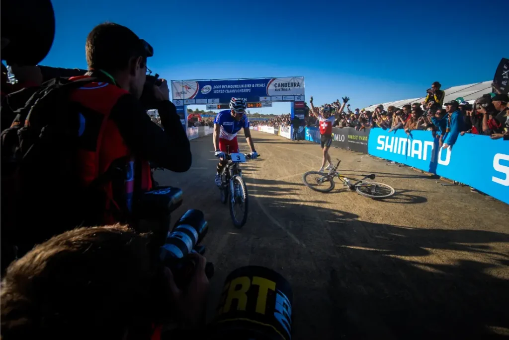 Un ciclista cruza la línea de meta mientras otro -posiblemente en señal de retirada de Nino Schurter- yace en el suelo, rodeado de fotógrafos y espectadores.