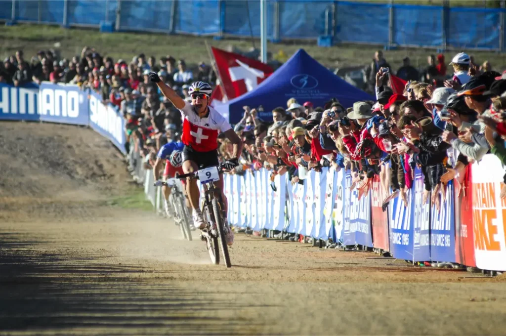 Un ciclista con un maillot suizo -que celebra la retirada de Nino Schurter- levanta un brazo en señal de victoria mientras cruza la línea de meta, animado por una multitud detrás de unas barricadas en una prueba ciclista.