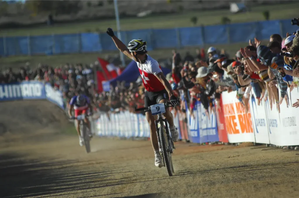 Un ciclista ataviado con un maillot blanco y rojo levanta un brazo en señal de victoria al cruzar la línea de meta, haciéndose eco de momentos como la retirada de Nino Schurter, con espectadores animando a lo largo del recorrido.