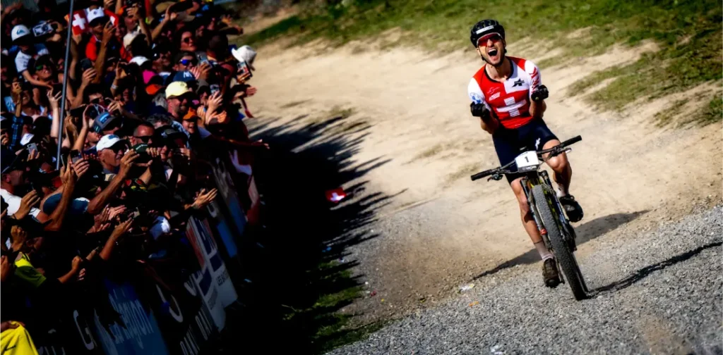 Un ciclista con maillot rojo y negro celebra al cruzar la línea de meta, evocando recuerdos del retirado Nino Schurter, mientras una multitud de espectadores anima en el lado izquierdo de una pista de tierra.