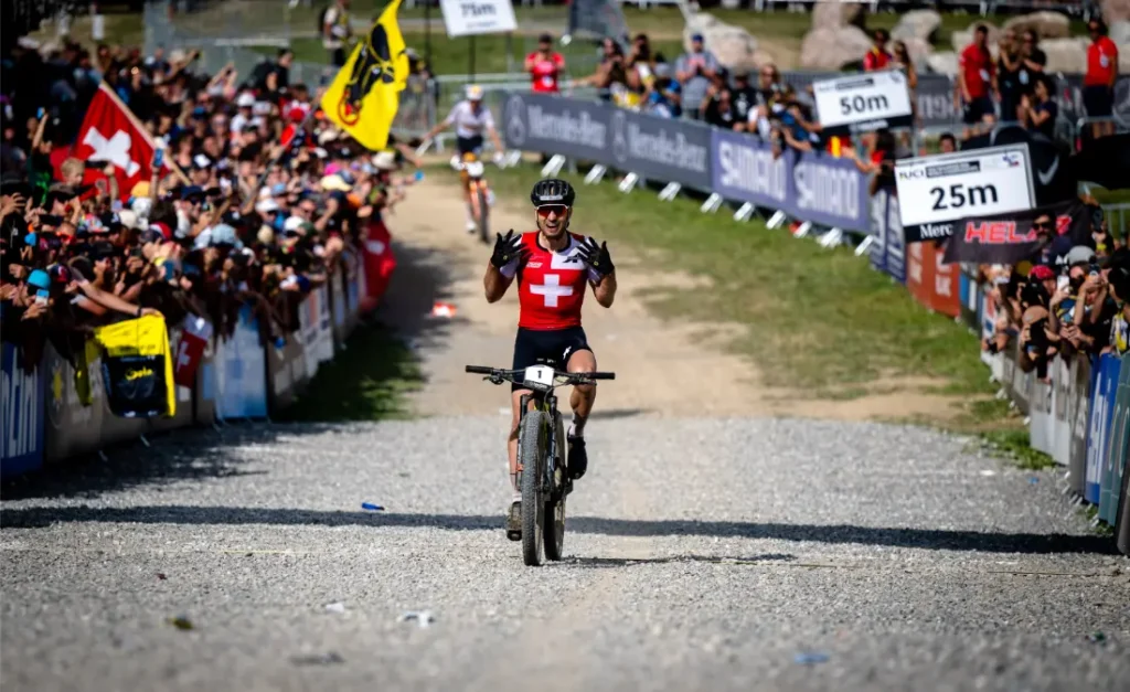 Un ciclista ataviado con un maillot rojo con una cruz blanca, que recuerda los momentos de retirada de Nino Schurter, celebra mientras cruza la línea de meta ante una multitud que le aclama en una carrera de bicicleta de montaña.