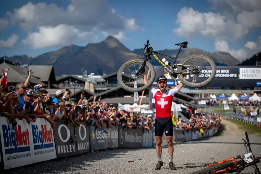 Un ciclista con maillot suizo, inspirado en el retirado Nino Schurter, levanta su bicicleta por encima de la cabeza en señal de celebración en una prueba de ciclismo de montaña, con espectadores animando y montañas de fondo.