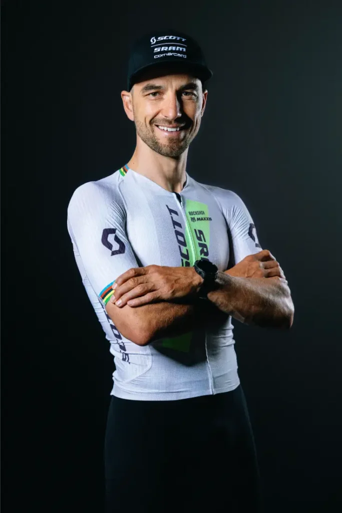 Un ciclista con un maillot blanco con los logotipos de los patrocinadores, de pie, con los brazos cruzados, sonriendo, y con una gorra negra sobre un fondo oscuro, captura el momento de la retirada de Nino Schurter.