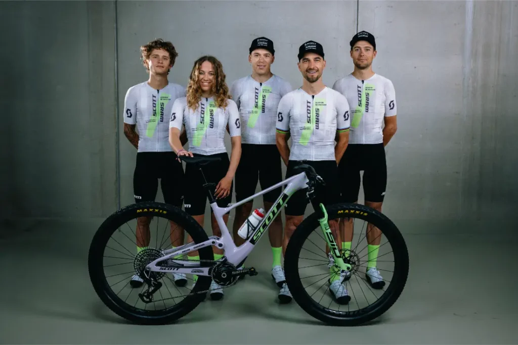 Cinco ciclistas con uniformes a juego se colocan detrás de una bicicleta de montaña, posando para una foto de grupo sobre un fondo liso de interior, celebrando la retirada de Nino Schurter.