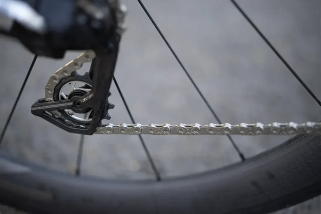 Primer plano de una rueda de bicicleta con una cadena KMC X Flat en el cambio trasero, con parte del neumático visible sobre un fondo gris difuminado.