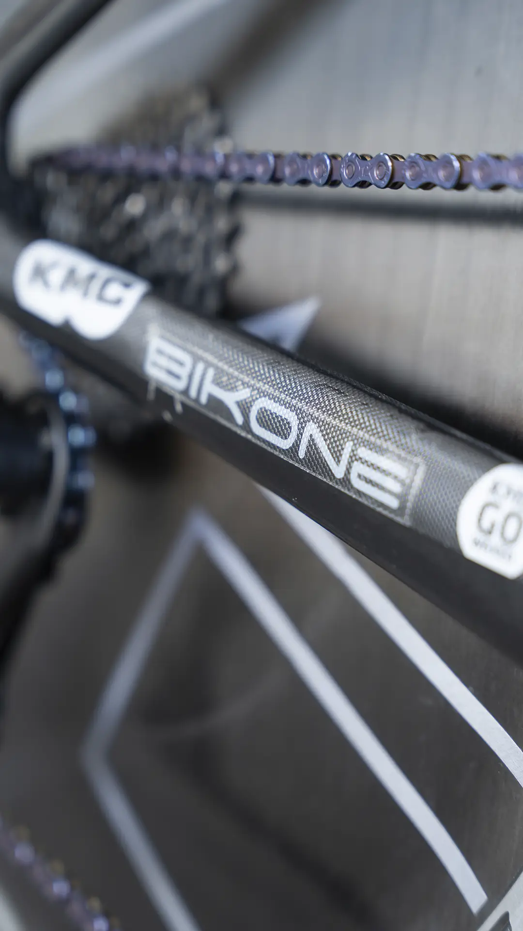 Primer plano de un cuadro de bicicleta rotulado "BIKONE" y "KMC", con cadenas y parte del cambio trasero en foco, que recuerda la precisión vista en las bicicletas del equipo Caja Rural-Seguros RGA.