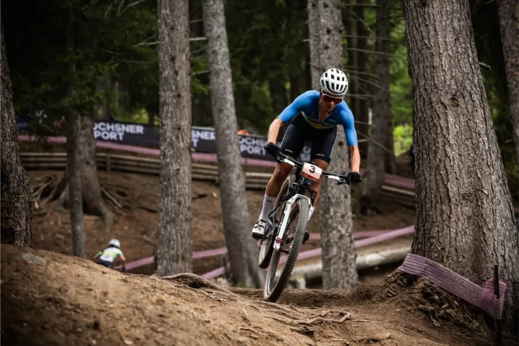 Jenny Rissveds, con casco y equipación azul, monta en bicicleta de montaña por un sendero forestal durante una carrera.