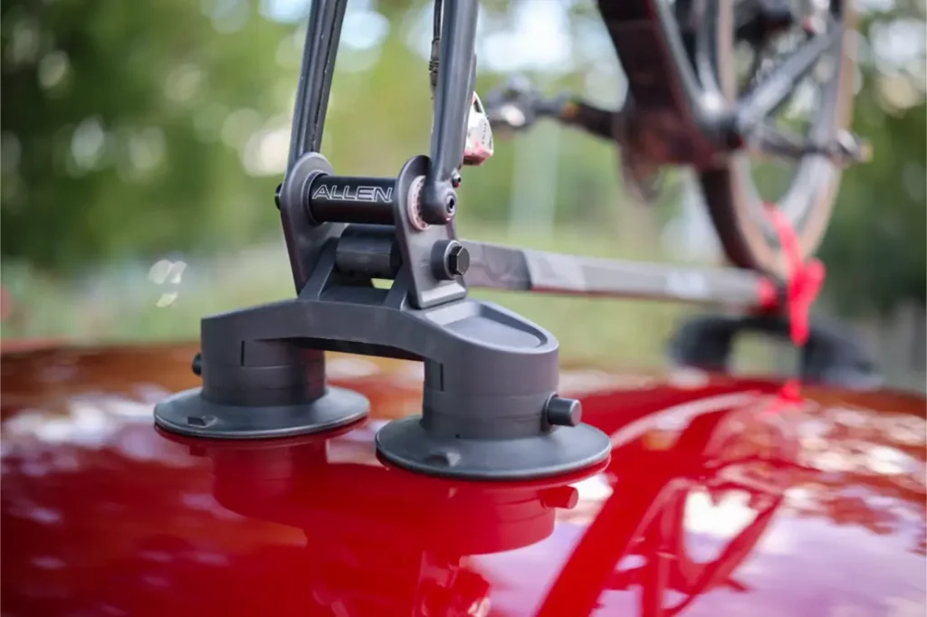 Primer plano de un portabicicletas Allen Smart Suction Bike Rack montado en el techo de un coche rojo brillante, sujetando parte de una bicicleta. Los árboles y la vegetación se difuminan en el fondo.