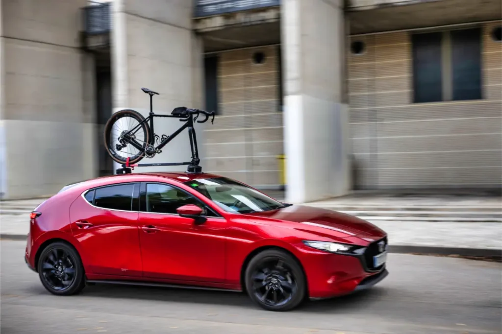 Un utilitario rojo con un portabicicletas Allen Smart Suction montado en el techo transporta una bicicleta mientras pasa por delante de un moderno edificio de hormigón.
