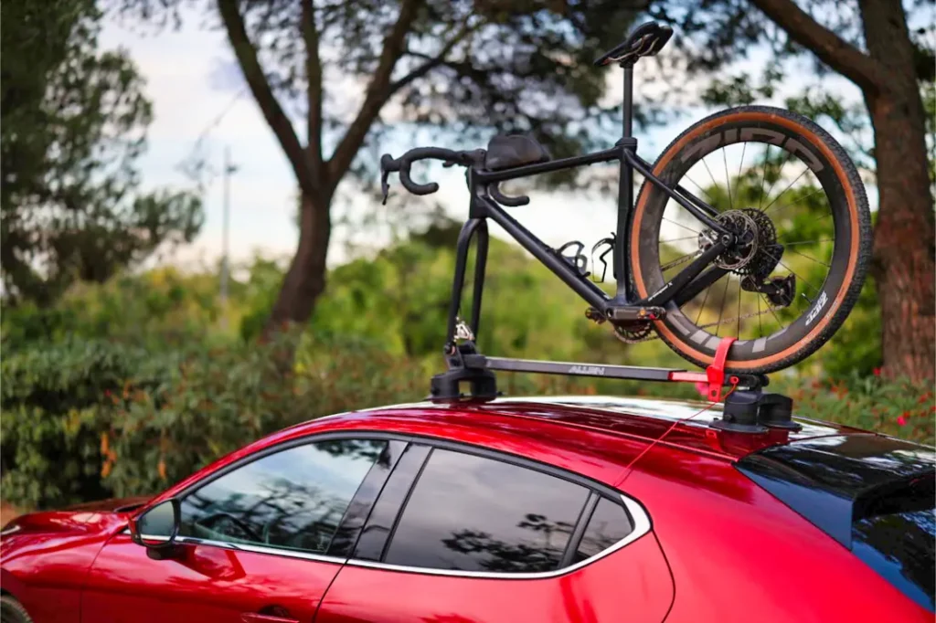 Una bicicleta está montada en un portabicicletas Allen Smart Suction Bike Rack sobre un coche rojo, aparcado al aire libre con árboles y vegetación de fondo.