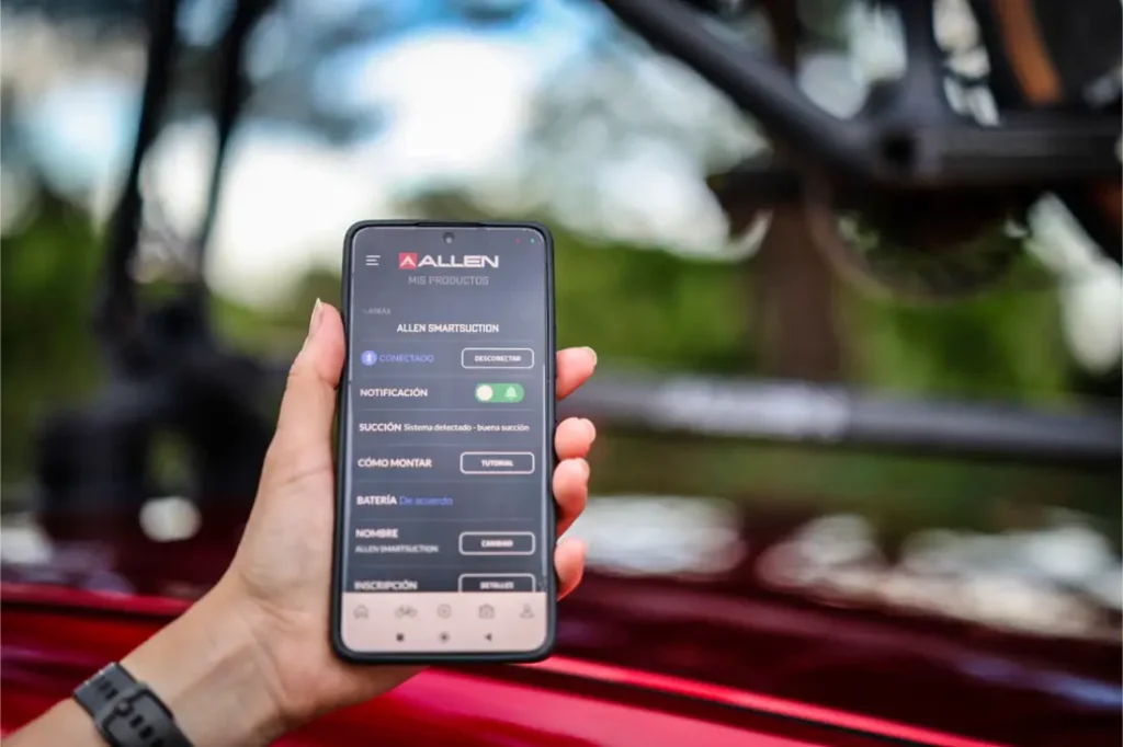 Una mano sostiene un smartphone que muestra la interfaz de la aplicación de Allen en español, con un fondo exterior difuminado y parte de una bicicleta equipada con Allen Smart Suction Bike Rack visible.