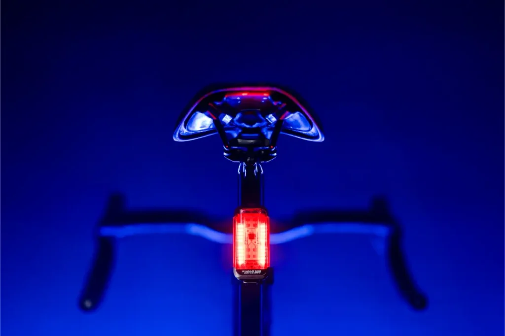 Bicicleta fotografiada desde atrás con una luz trasera Lezyne Radar React System iluminada bajo el sillín, manillar visible, sobre fondo azul.