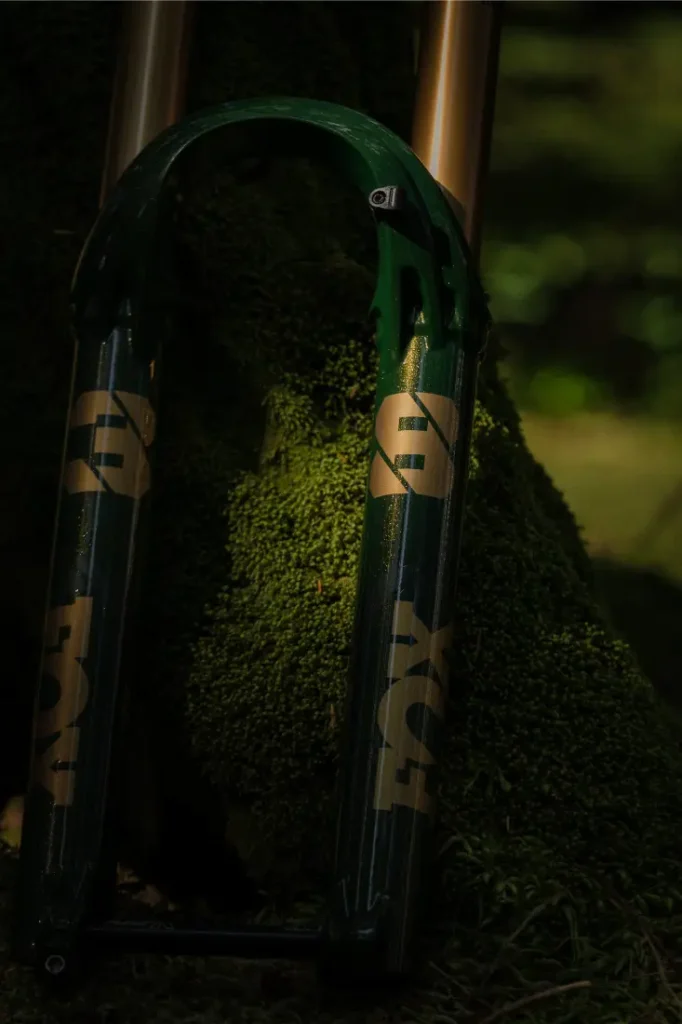 Primer plano de una horquilla de suspensión de bicicleta FOX 38 Racing Green Edición Limitada apoyada en un árbol cubierto de musgo en un entorno forestal sereno.