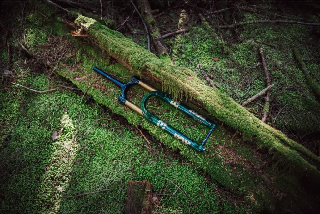 Una horquilla de suspensión FOX 38 Racing Green Edición Limitada con un llamativo acabado verde y dorado descansa sobre un tronco cubierto de musgo en un apacible entorno forestal.