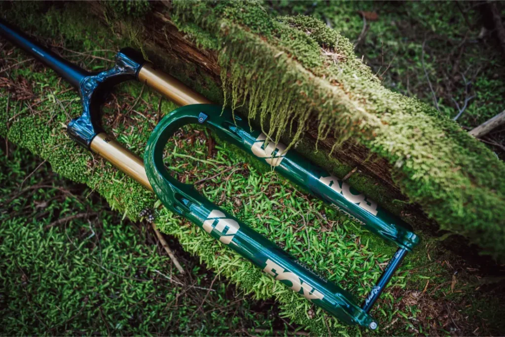 Una horquilla de suspensión FOX 38 Racing Green Edición Limitada en verde vibrante y dorado descansa sobre un suelo musgoso junto a un tronco cubierto de musgo.