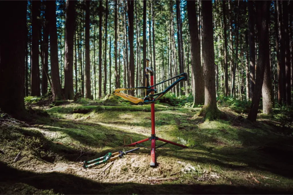 Un cuadro de bicicleta de montaña, con la horquilla FOX 38 Racing Green, está montado en un puesto de reparación en un bosque iluminado por el sol, con herramientas y piezas de bicicleta esparcidas por el musgoso suelo cercano.