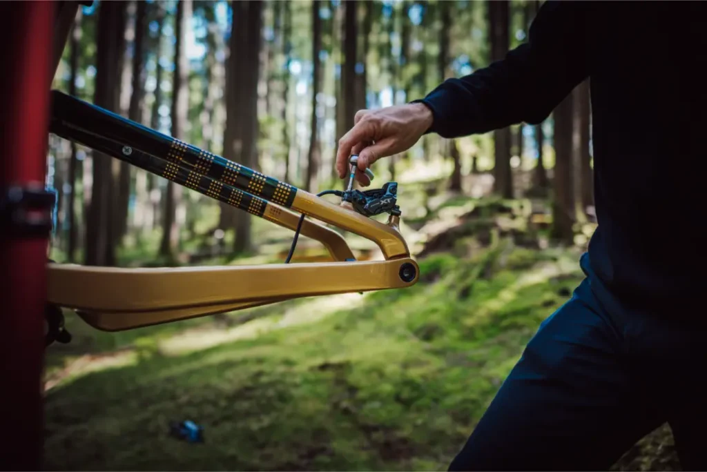 Una persona ajusta el cambio trasero de una bicicleta de montaña amarilla equipada con un FOX 38 Racing Green Edición Limitada en un sereno entorno forestal.