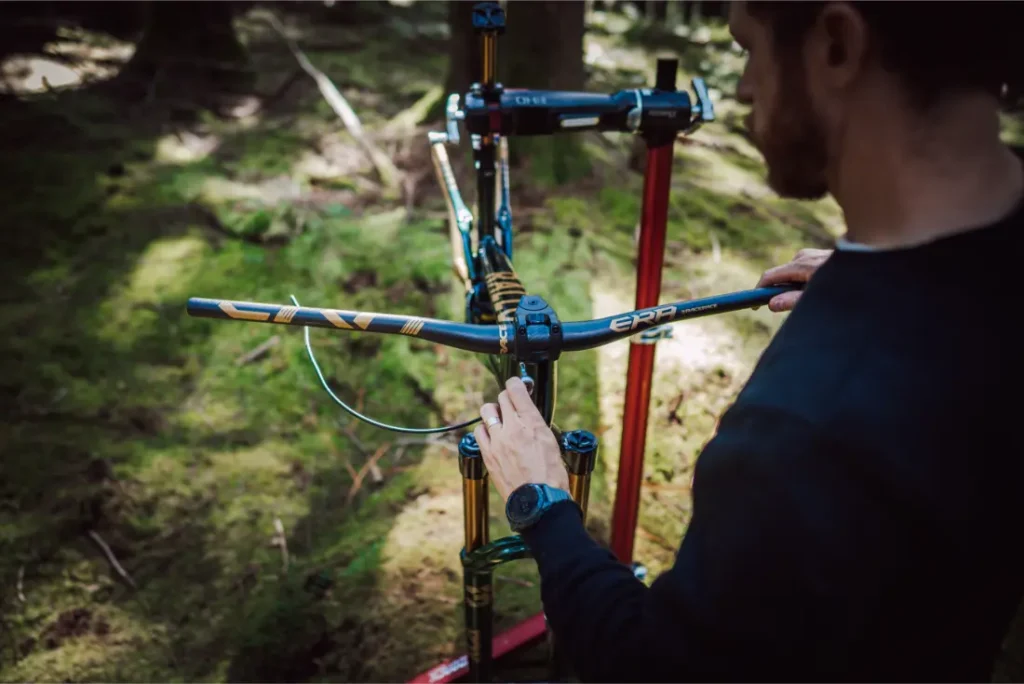 Una persona ajusta el manillar de una bicicleta de montaña con horquilla FOX 38 Racing Green Edición Limitada, montada en un soporte de reparación en una zona boscosa.