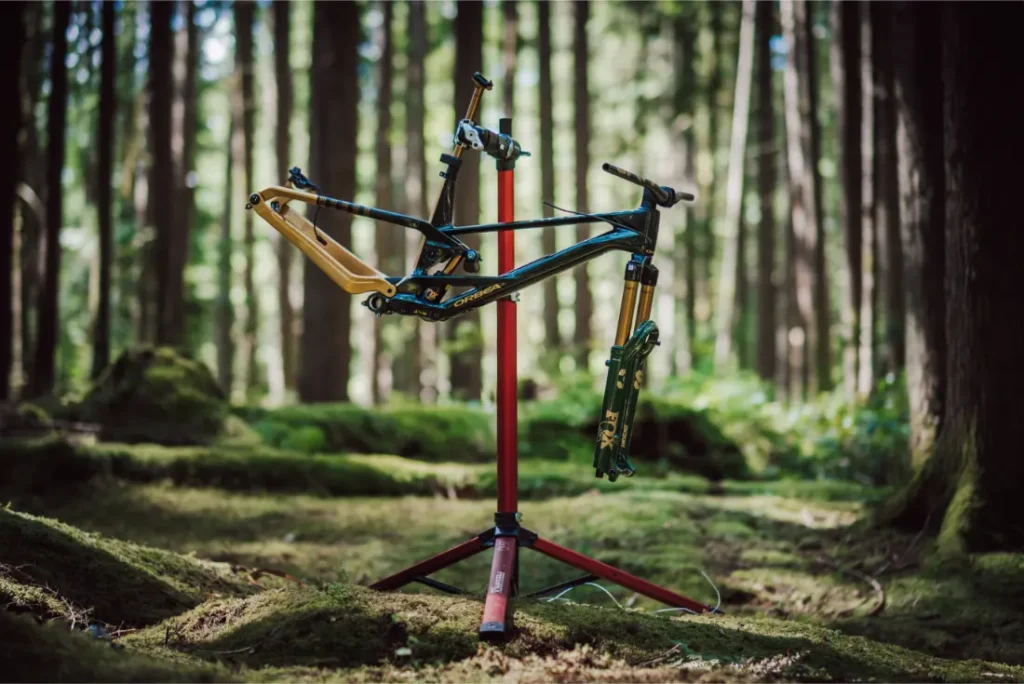 Un cuadro de bicicleta de montaña parcialmente montado con una horquilla FOX 38 Racing Green montada en un soporte de reparación en un entorno de bosque musgoso, con altos árboles alzándose al fondo.