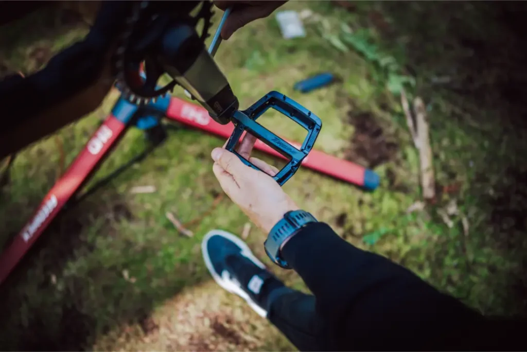 Una persona instala un pedal de bicicleta azul en un brazo de biela al aire libre, con césped y un soporte de bicicleta rojo visibles en el fondo, mientras una horquilla FOX 38 Racing Green Edición Limitada llama la atención.