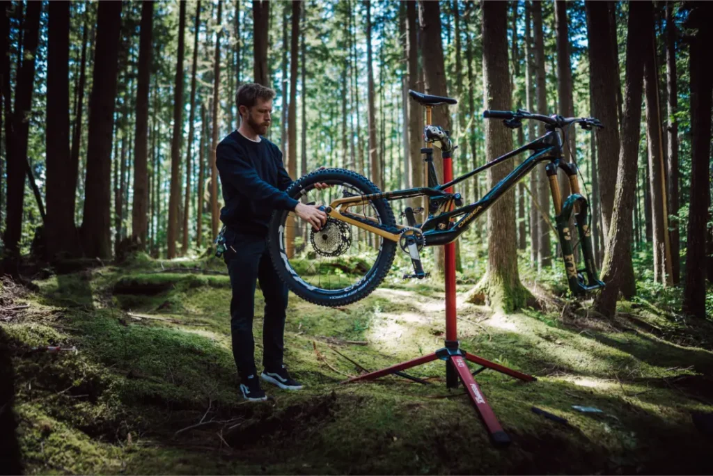 Una persona ajusta una bicicleta de montaña con un FOX 38 Racing Green Edición Limitada en un puesto de reparación en un bosque, rodeado de altos árboles y la luz del sol filtrándose a través del dosel.