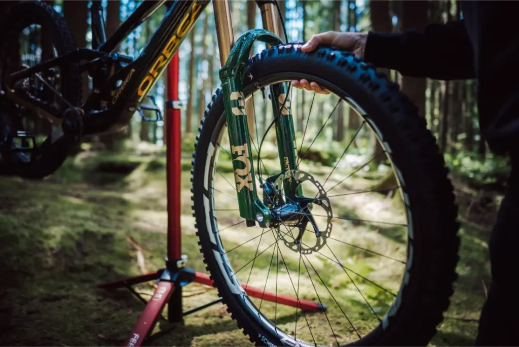 Una persona instala una rueda delantera en una bicicleta de montaña equipada con una suspensión FOX 38 Racing Green Edición Limitada en una zona boscosa, utilizando un soporte de bicicleta rojo como apoyo.
