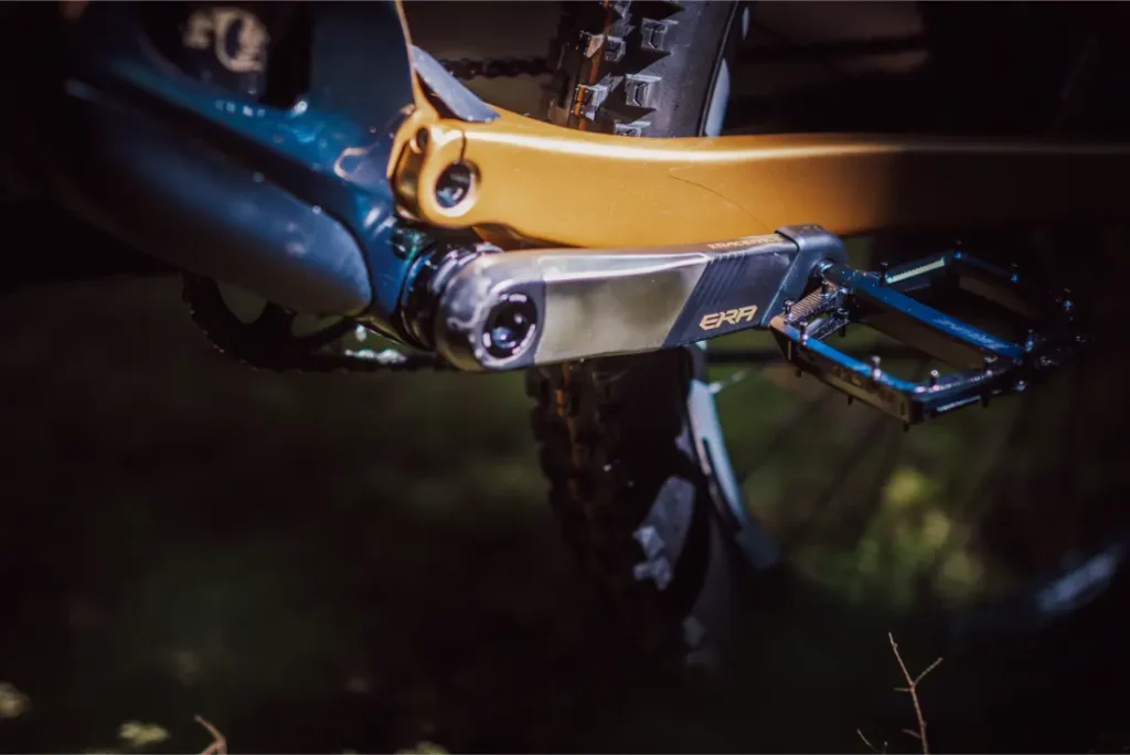 Primer plano de un brazo de biela de bicicleta con la etiqueta "EXR" y pedal plano fijado al cuadro naranja y azul, acentuado por la horquilla FOX 38 Racing Green, con parte de la rueda trasera y la cadena visibles.