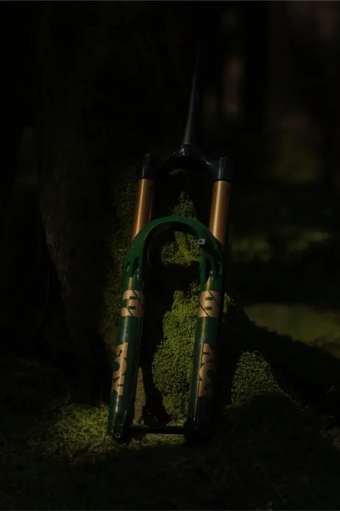 Una horquilla de suspensión de bicicleta de montaña FOX 38 Racing Green está apoyada contra un árbol musgoso en un bosque poco iluminado, mostrando su llamativo acabado verde y dorado Edición Limitada.