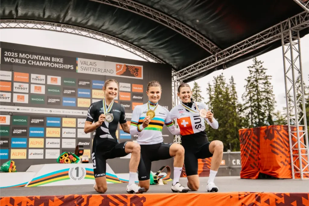 Tres ciclistas, entre ellas Jenny Rissveds, posan en el podio con sus medallas tras un campeonato de ciclismo de montaña; al fondo se ven los logotipos de los patrocinadores y las pancartas del evento.