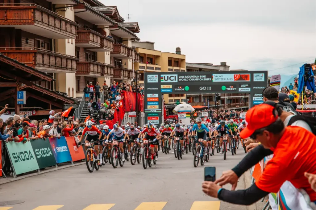 Ciclistas compiten en los Campeonatos del Mundo de Bicicleta de Montaña de la UCI de 2025 en Valais, Suiza, mientras el público observa cerca de la línea de salida, aclamando a estrellas como Jenny Rissveds.