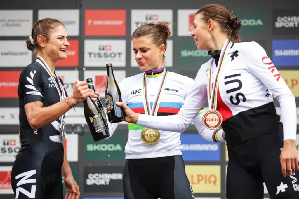Tres ciclistas, entre ellas Jenny Rissveds, con medallas, de pie en un podio, sosteniendo botellas y celebrando frente a un telón de fondo de patrocinadores.