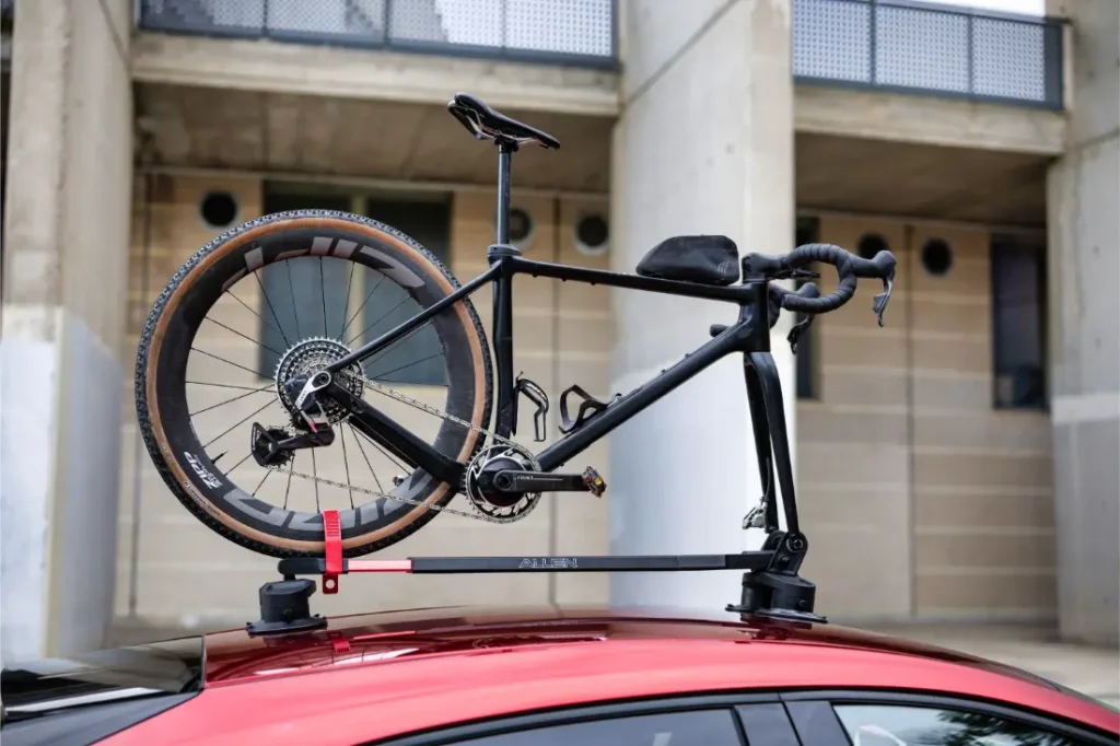 Una bicicleta de carretera negra montada boca abajo en un portabicicletas Allen Smart Suction Bike Rack sobre un coche rojo, con un edificio de hormigón al fondo.