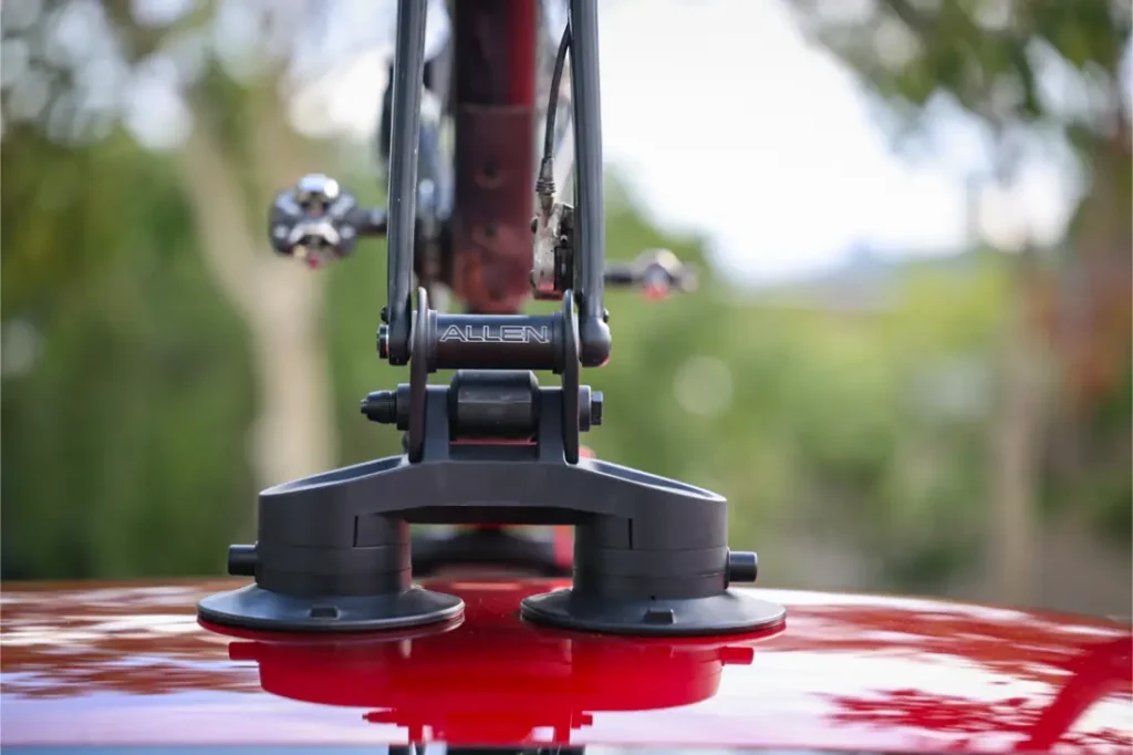 Primer plano de un portabicicletas Allen Smart Suction montado en el techo de un coche rojo, con la marca "Allen" visible. Los árboles aparecen difuminados en el fondo, resaltando el elegante diseño de este portabicicletas Allen Smart Suction.