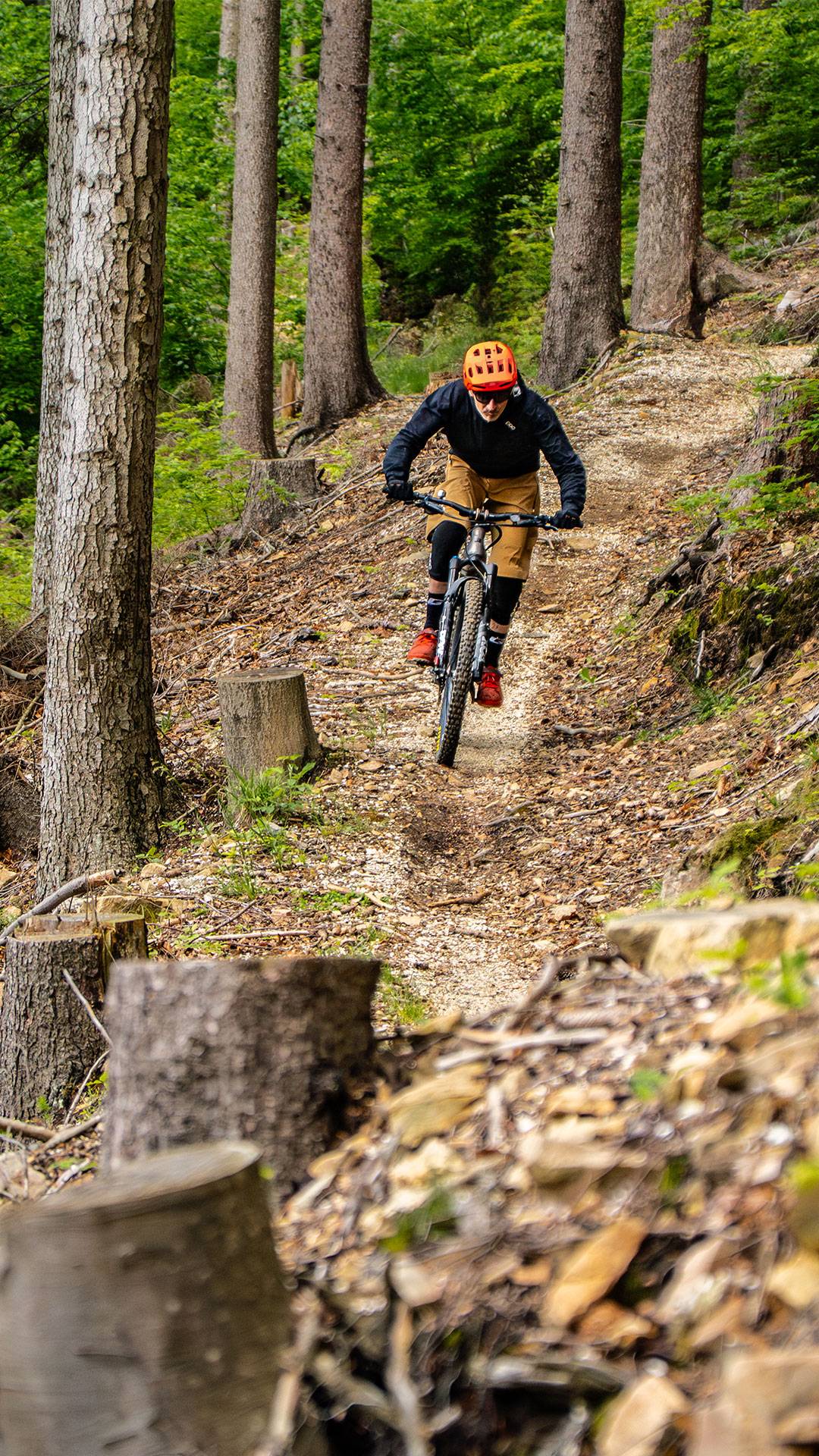 Persona con casco naranja y zapatos rojos monta en bicicleta de montaña por un estrecho sendero de tierra entre árboles altos y follaje verde en el pintoresco bosque Bike Region Glacensis.