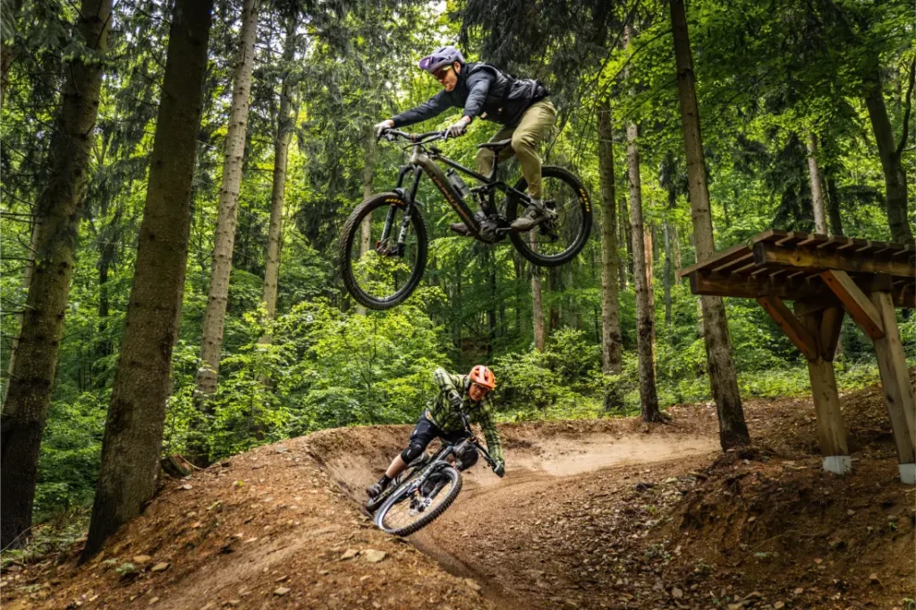 Dos ciclistas de montaña recorren un sendero forestal en la pintoresca Bike Region Glacensis; uno salta desde una rampa mientras el otro toma una curva de tierra más abajo.