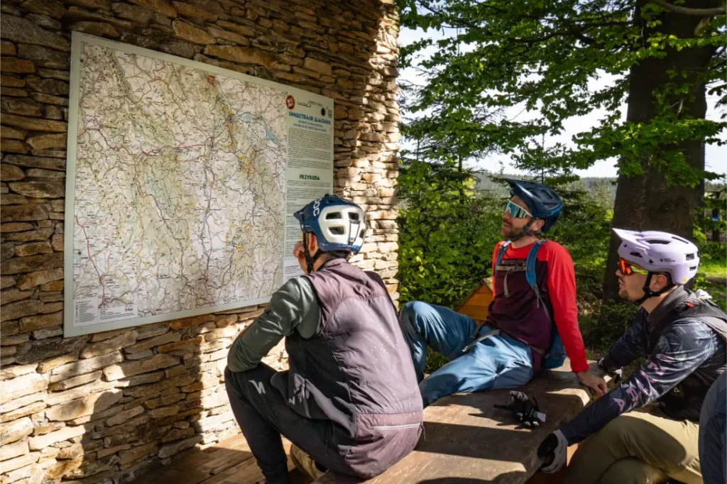 Tres ciclistas con casco se sientan y estudian un gran mapa exterior de la Bike Region Glacensis, montado en un muro de piedra, con vegetación visible al fondo.