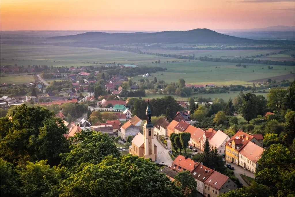 Un pequeño pueblo con edificios de tejados rojos y una iglesia está rodeado de campos verdes y árboles, rodeado de colinas onduladas al atardecer en la pintoresca Bike Region Glacensis.