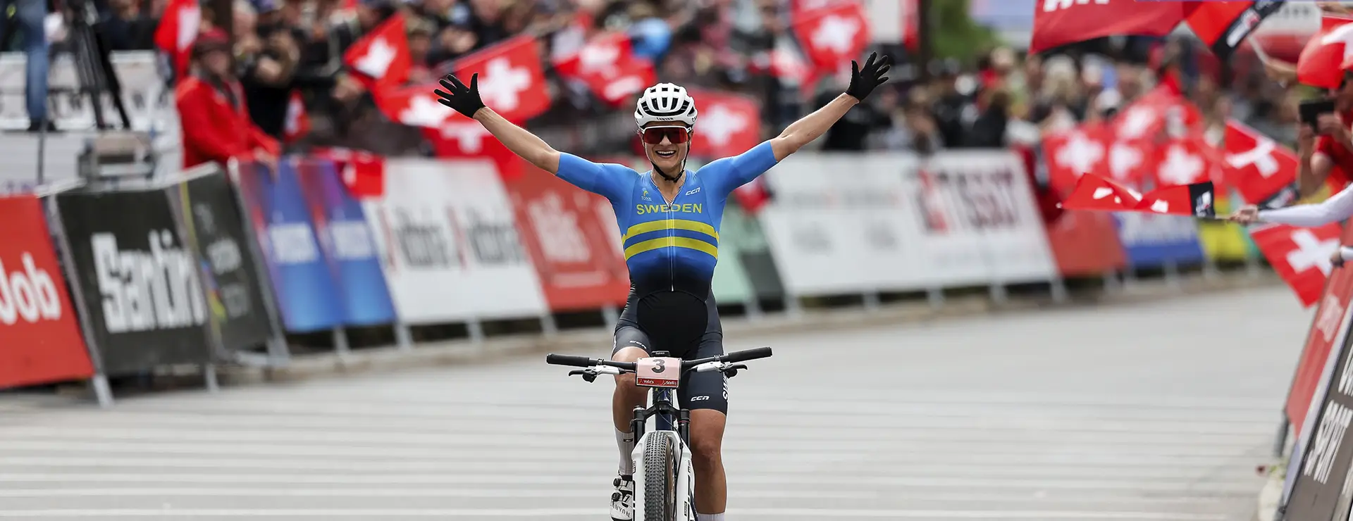Una ciclista con el maillot de Suecia -Jenny Rissveds, oro en Río- levanta ambos brazos en señal de celebración al cruzar la línea de meta, con espectadores ondeando banderas suizas al fondo, captando su cima del mundo.