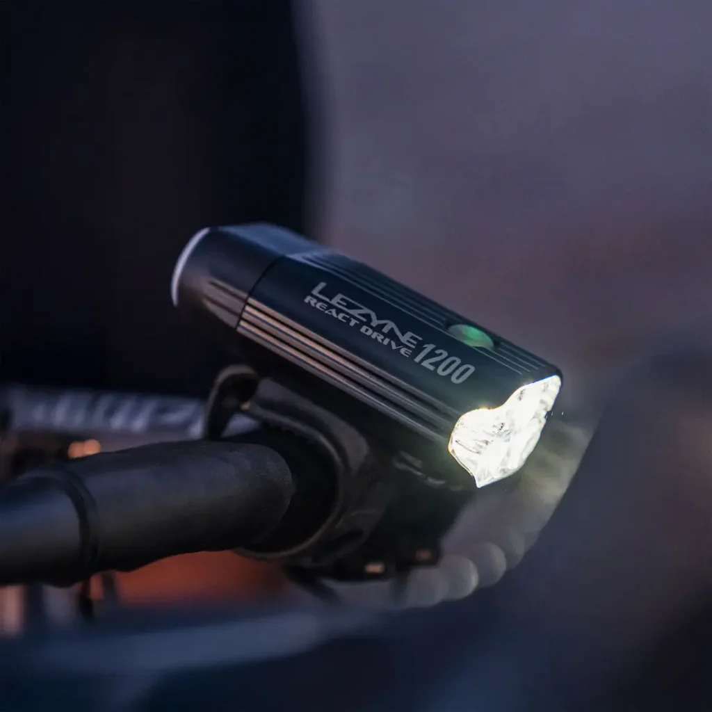 Un faro para bicicleta Lezyne React Drive 1200 con sistema Lezyne Radar React, montado en el manillar e iluminado en condiciones de poca luz.