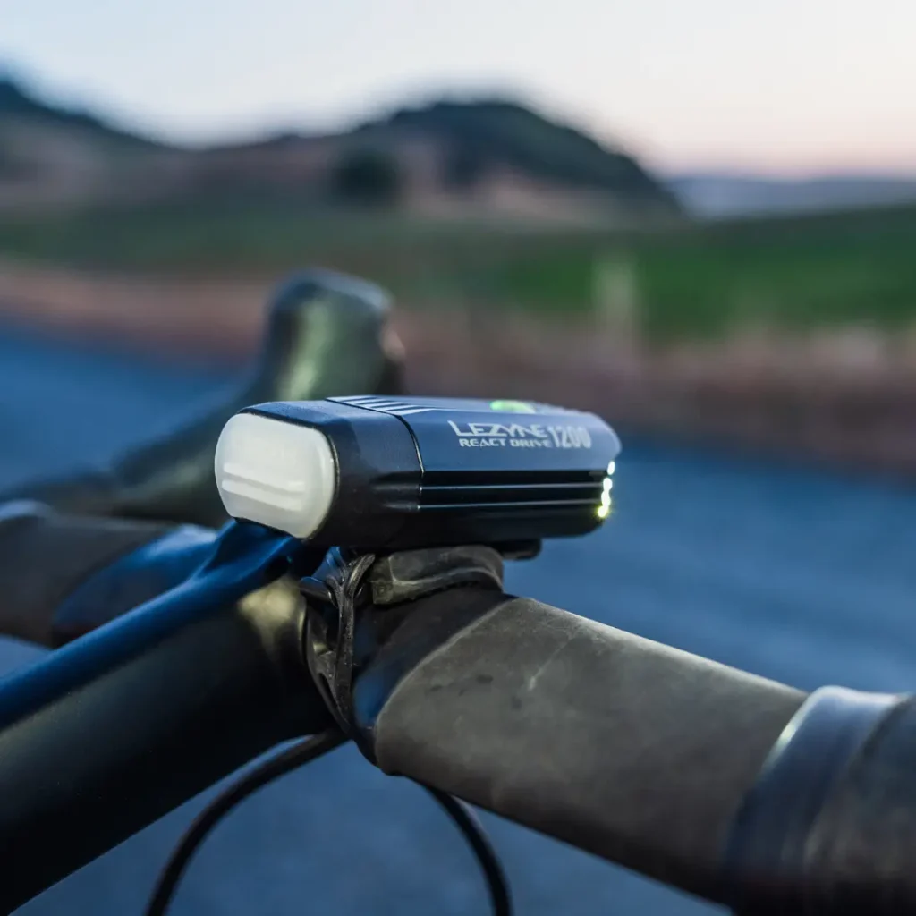 Primer plano de una luz de bicicleta Lezyne React Drive 1200 con sistema Lezyne Radar React montada en el manillar de una bicicleta, con un paisaje exterior borroso de fondo.