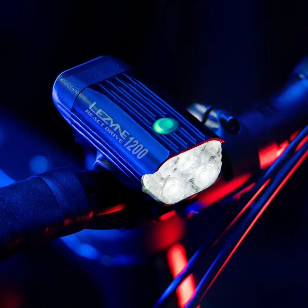 Un faro para bicicleta Lezyne React Drive 1200, que incorpora el avanzado sistema Lezyne Radar React, está montado en el manillar e iluminado sobre un fondo oscuro con detalles luminosos en azul y rojo.