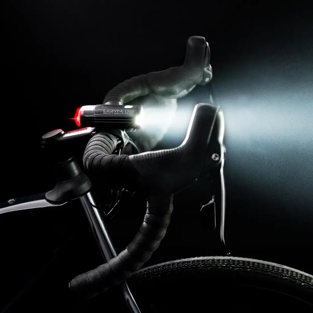 Primer plano del manillar y la rueda delantera de una bicicleta en la oscuridad, con un brillante faro Lezyne Radar React System acoplado al manillar e iluminando la escena.