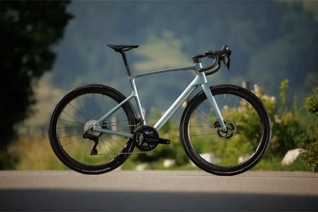 Una bicicleta de carretera SCOTT Addict con manillar rebajado, frenos de disco y neumáticos finos está aparcada en una superficie pavimentada con vegetación y colinas al fondo.
