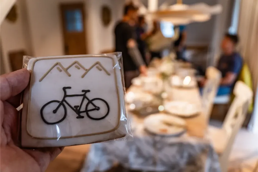 Una mano sostiene una galleta cuadrada decorada con una bicicleta y montañas cerca de una mesa de comedor preparada para una comida, capturando el espíritu de Bike Region Glacensis con gente reunida al fondo.