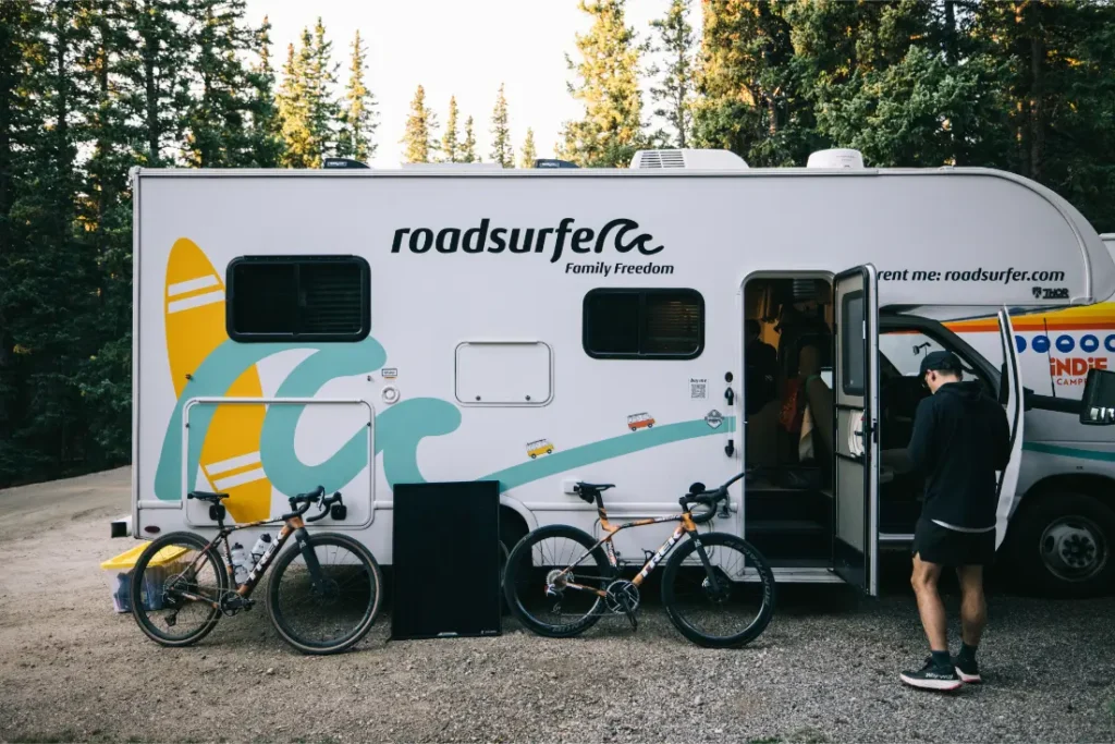 Un hombre junto a dos bicicletas aparcadas delante de una furgoneta camper "roadsurfer" en una zona boscosa, evocando el espíritu de aventura que se encuentra en States of Elevation - Kilian Jornet.
